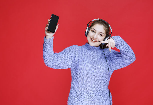 服装戴着耳机的女孩展示着她的智能手机 感觉很开心满意积极促销