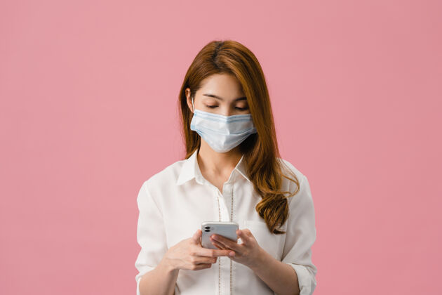 感染年轻的亚洲女孩戴着医用面罩用手机 穿着粉色背景隔离的休闲服卫生流行病隔离