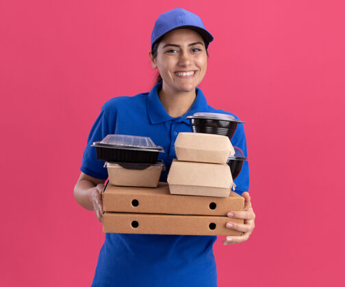 女孩带着微笑的年轻送货女孩 穿着制服 戴着帽子 把食物容器放在隔离在粉红色墙上的比萨饼盒上穿着人表情