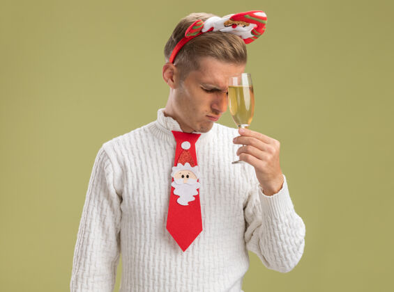 橄榄紧张的年轻帅哥戴着圣诞老人的头带打着领带拿着香槟酒杯抚摸着额头闭着眼睛隔离在橄榄绿的墙上留着复印空间领带圣诞老人年轻