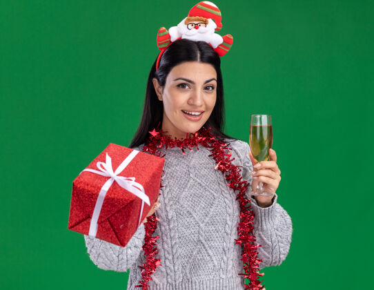 周围年轻的白人女孩戴着圣诞老人的头带 脖子上戴着金箔花环 手里拿着一杯香槟 向绿色的墙上伸出礼包花环圣诞老人圣诞老人