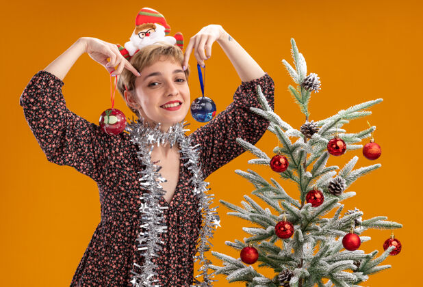 立场微笑着的年轻漂亮女孩戴着圣诞老人的头带 脖子上戴着金箔花环 站在装饰好的圣诞树旁 拿着圣诞饰品 靠近孤立在橙色墙上的头微笑近头