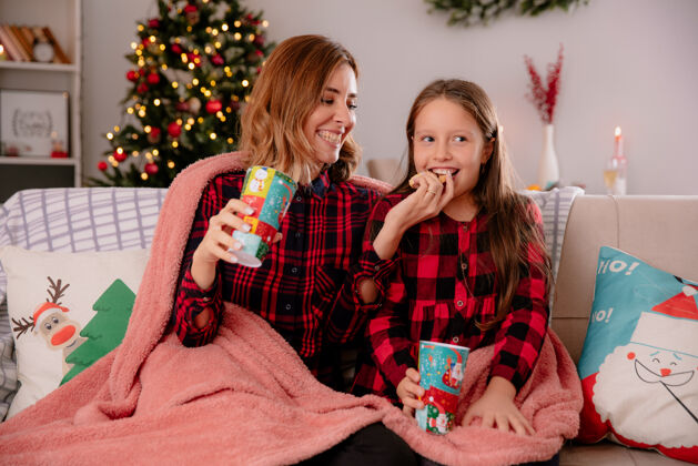沙发微笑的母亲拿着纸杯 拿着饼干喂女儿 坐在铺着毯子的沙发上 在家里享受圣诞节时光女儿享受抱