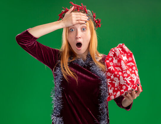 圣诞节年轻漂亮的女孩穿着红色的裙子 脖子上戴着花环和花环 手里拿着圣诞包 手放在额头上 被隔离在绿色的墙上害怕穿脖子