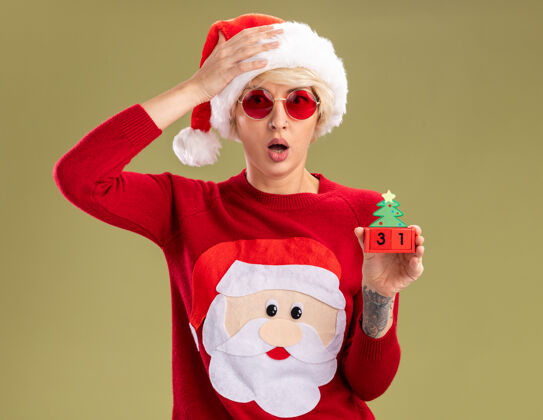 保持戴着圣诞帽 戴着圣诞眼镜 穿着圣诞毛衣 手抱着圣诞树玩具 看起来像个约会的金发女郎 手放在头上 隔离在橄榄绿的墙上圣诞节手穿