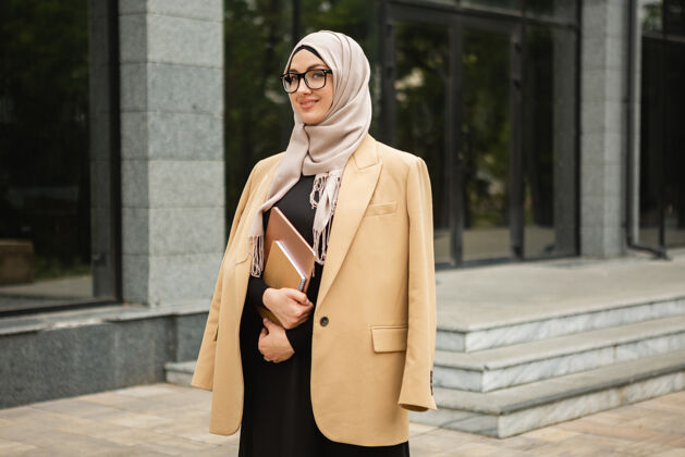 女性现代时尚的妇女戴着头巾 穿着商务风格的夹克和黑色的长袍 带着笔记本电脑走在城市街道上自信头巾伊斯兰