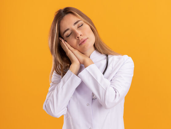 衣服年轻的女医生闭着眼睛 穿着医用长袍 听诊器显示睡眠姿势 隔离在黄色的墙上表情感觉封闭