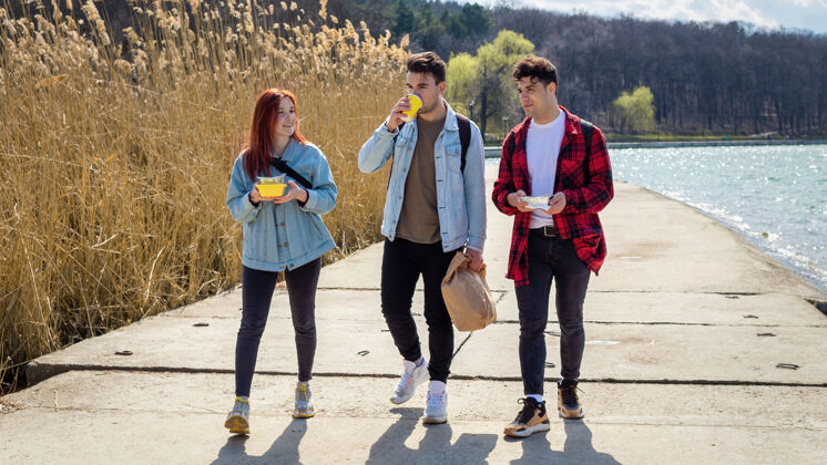 年轻三个年轻的朋友在湖边散步 在公园里吃喝团体放松公园