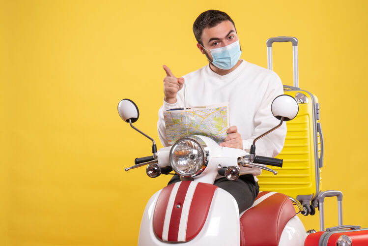 人旅行概念的俯视图 戴着医用面罩的自信男子站在摩托车旁 黄色手提箱放在上面 拿着地图指向前方视图地图旅行
