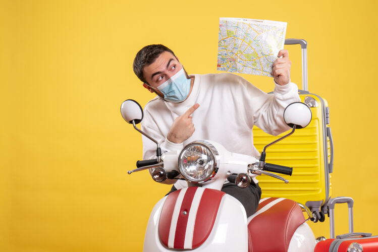 面罩旅行概念的俯视图 戴着医用面罩的好奇的家伙站在摩托车旁边 黄色的手提箱在上面 指着地图人站立球
