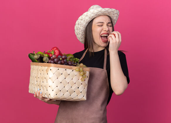 不愉快漂亮的白人女园丁戴着园艺帽 拿着菜篮子 假装咬西红柿粉色抱花园