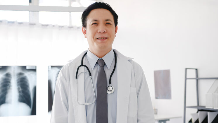 医疗年轻的亚洲男医生穿着白色的医疗制服 手持听诊器看着摄像机 微笑着双臂交叉着 在卫生院与患者进行视频电话会议疾病看着摄像机听诊器