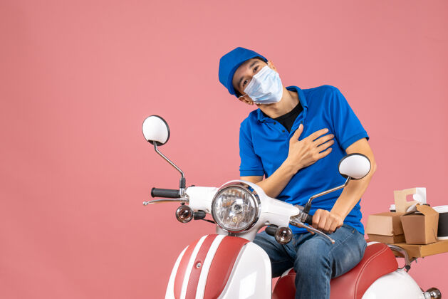 摩托车正面图：戴着医疗面罩 戴着帽子 坐在粉彩桃色背景下的踏板车上的快递员背景头盔粉彩
