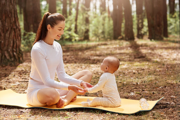 童年侧视图快乐年轻的运动母亲坐在卡雷马特在森林与她的婴儿家庭产前婴儿