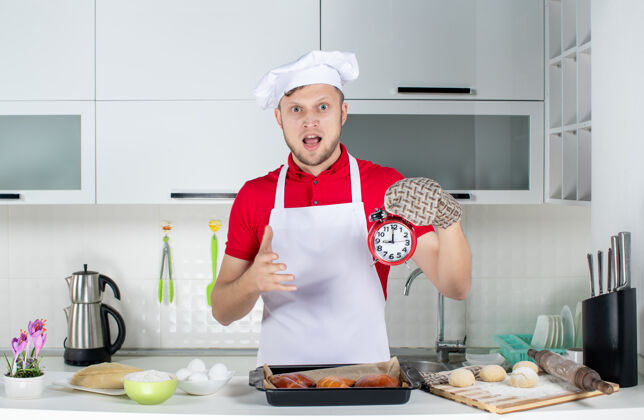 年轻在白色厨房里 年轻惊讶的男厨师戴着支架拿着钟 做着ok的手势国内支架时钟