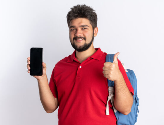 站立年轻的留着胡子的学生 穿着红色马球衫 背着背包 露出智能手机 微笑着 自信地竖起大拇指 站在白色的墙上拇指表演年轻人