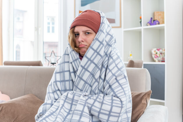 流感不健康的年轻女子戴着帽子 裹着毯子 感觉不舒服 生病 患感冒和流感 发烧 坐在明亮的客厅沙发上女人发烧光