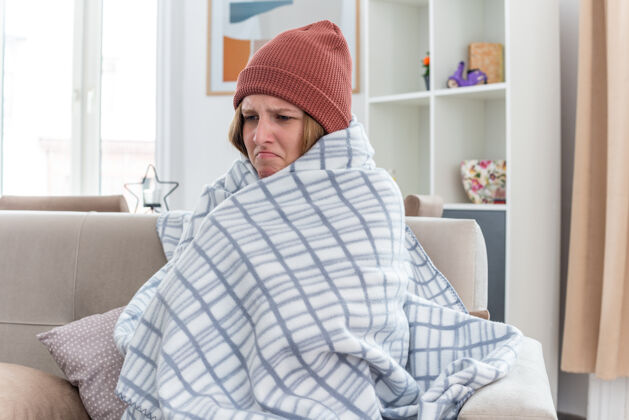 帽子不健康的年轻女子 戴着暖和的帽子 裹着毯子 看起来不舒服 生病 感冒 流感 发烧 头痛 坐在客厅里的沙发上女人温暖不适