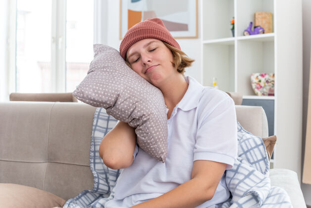 毯子微笑不健康的年轻女子 戴着暖和的帽子 拿着毛毯 患感冒和流感 抱着枕头 头靠在枕头上 闭着眼睛 坐在明亮的客厅沙发上光精益头