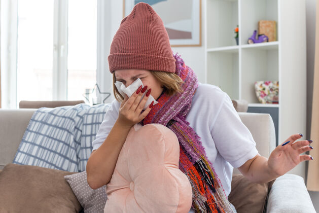 生活不健康的年轻女子戴着暖和的帽子 戴着围巾 抱着枕头 用纸巾擤鼻涕 患有感冒和流感 坐在明亮的客厅椅子上枕头流感女人