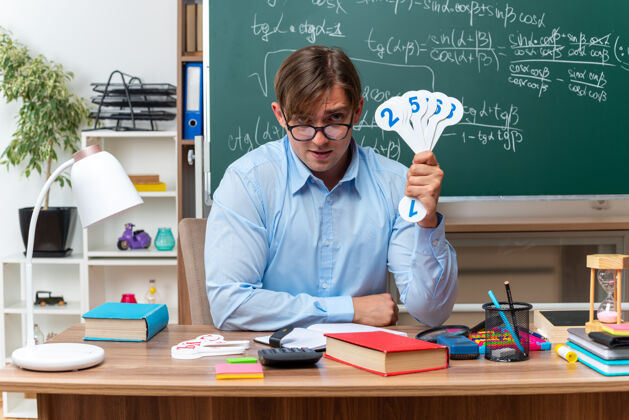 笔记年轻的男老师戴着眼镜 展示着解释课的车牌 微笑着坐在课桌旁 教室里的黑板前放着书和笔记黑板学校教室