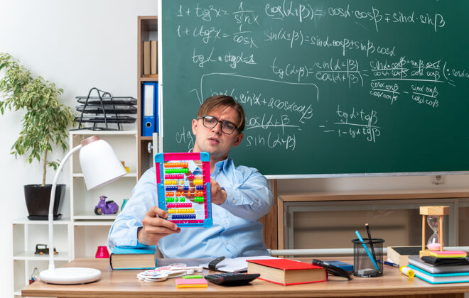 教室年轻的男老师戴着眼镜 带着钞票 看起来很自信 坐在课桌旁 在教室的黑板前放着书和笔记小笔记学校