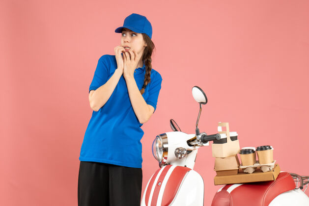 人困惑的女快递员站在摩托车旁边 咖啡和小蛋糕在粉彩桃色背景上成人粉彩人