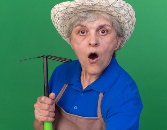 花园惊讶的上了年纪的女园丁戴着园艺帽拿着锄头耙看着相机绿色园艺穿