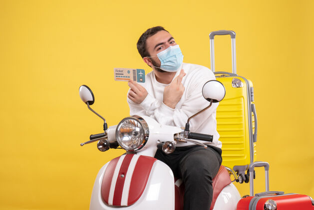 医疗旅行理念的俯视图 戴着医用面罩的充满希望的家伙坐在摩托车上 黄色手提箱在上面 手里拿着车票运动摩托车手持