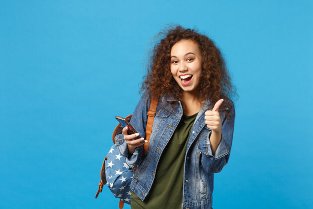 短信年轻的非洲裔美国女孩十几岁的学生在牛仔服装 背包举行电话隔离在蓝色的墙壁上电话手机女性