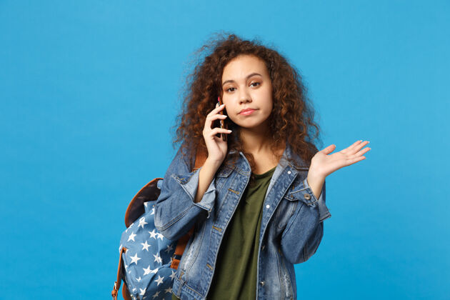 女人年轻的非洲裔美国女孩十几岁的学生在牛仔服装 背包举行电话隔离在蓝色的墙壁上电话沉思牛仔裤