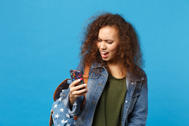 人年轻的非洲裔美国女孩十几岁的学生在牛仔服装 背包举行电话隔离在蓝色的墙壁上女孩手机设备