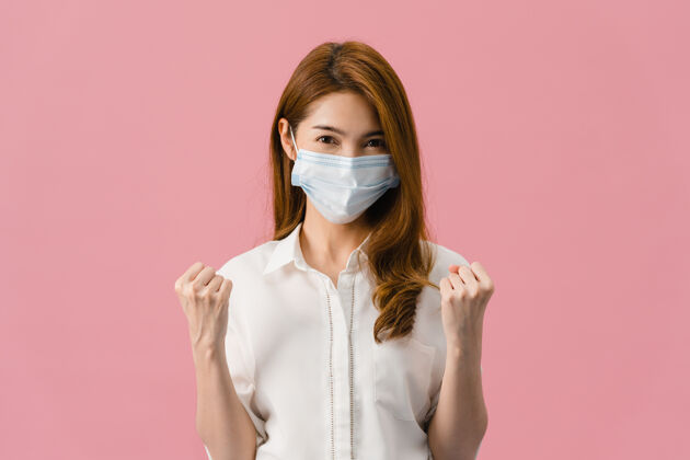 污染年轻的亚洲女孩戴着医用口罩显示和平的迹象 穿着休闲的衣服 看着隔离在粉红色背景上的相机鼓励患者疾病热情