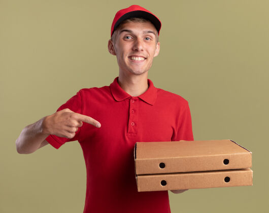 绿色微笑着的年轻金发送货员抱着并指着比萨饼盒点金发微笑