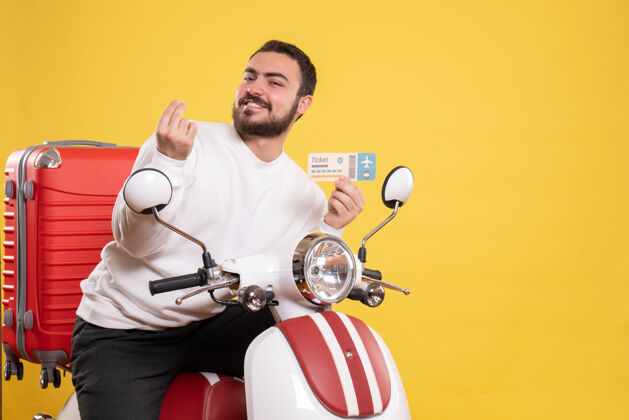手势正面图是一个微笑的年轻人坐在摩托车上 提着手提箱 在孤立的黄色背景下做着赚钱的手势票车辆高尔夫