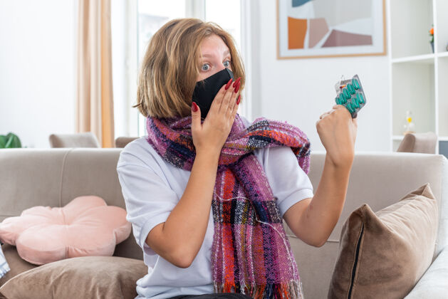 生活不健康的年轻女子 脖子上围着暖和的围巾 戴着面膜 感觉不舒服 生病 患流感 吃感冒药 坐在客厅的沙发上 看起来很困惑痛苦保护沙发