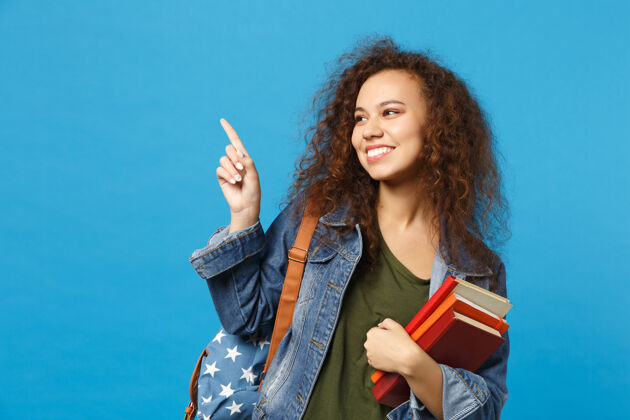 微笑穿着牛仔服 背着书包的年轻学生把书孤立地放在蓝色的墙上教学背校园