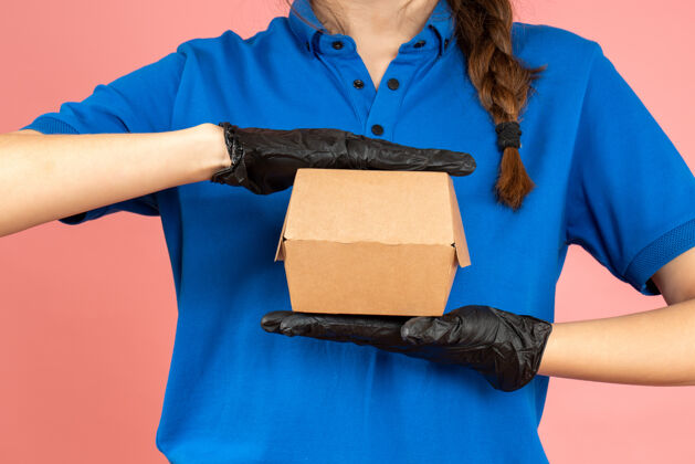 手持半身镜头的信使女孩戴着黑色手套拿着粉彩桃色背景上的小盒子射击防护服微笑