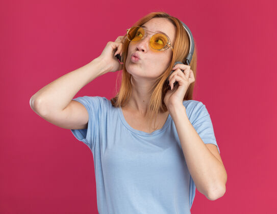 粉红色令人印象深刻的年轻红发姜女孩雀斑在太阳眼镜和耳机看一边姜眼镜印象