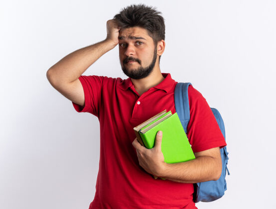 手势一个年轻的留着胡子的学生 穿着红色马球衫 背着背包 手里拿着笔记本 头上拿着一只手 因为他站在白墙上犯了个错误胡子错误困惑