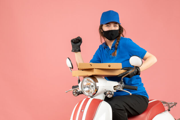 摩托车前视图年轻的快乐微笑的女快递员戴着医用面罩和手套坐在滑板车上传递粉彩桃背景的命令前面面具交付