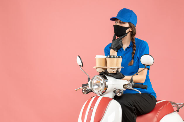 桃前视图体贴的女性送货员戴着医用面罩和手套坐在滑板车上拿着粉彩桃色背景的订单穿着头盔女