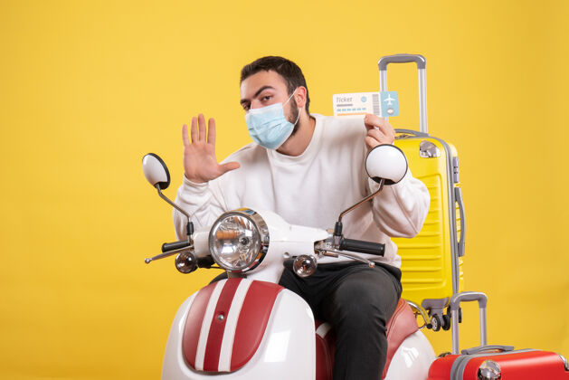 面具旅行概念的俯视图 戴着医用口罩的年轻人坐在摩托车上 黄色手提箱在上面 手里拿着五张票年轻人坐运动