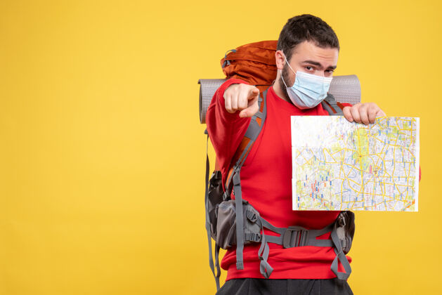 黄色顶视图年轻自信的旅行者家伙戴着医用面罩 背包里拿着黄色的地图指向前方人面具脸