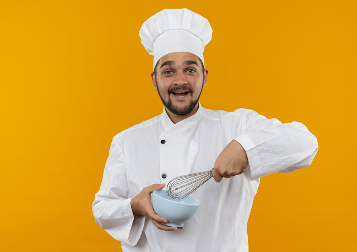 男快乐的年轻男厨师穿着厨师制服 拿着搅拌器和碗 隔离在橙色的墙上 留有复制空间制服碗拿着