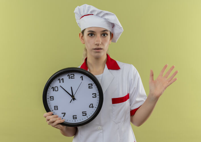 显示年轻漂亮的厨师穿着厨师制服拿着时钟 在绿色的墙上显示出孤立的空手印象深刻钟手厨师