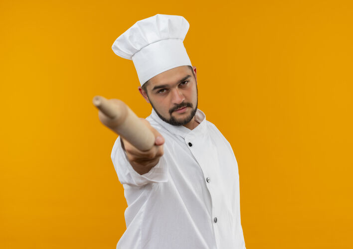 男性自信的年轻男厨师身着厨师制服 将擀面杖伸向孤立的橙色墙壁 留有复制空间别针伸展滚动