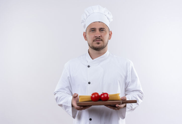 烹饪自信的年轻帅哥厨师身着厨师制服 手拿着砧板 上面有西红柿和意大利面 隔离在白色的墙上年轻持有意大利面
