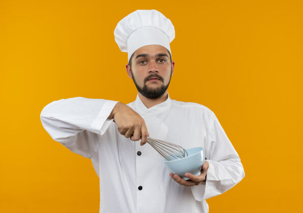 年轻令人印象深刻的年轻男性厨师在厨师制服拿着威士忌和碗隔离在橙色墙上搅拌印象厨师
