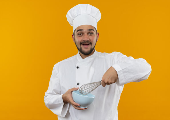 男快乐的年轻男厨师穿着厨师制服 拿着搅拌器和碗 隔离在橙色的墙上 留有复制空间制服碗拿着